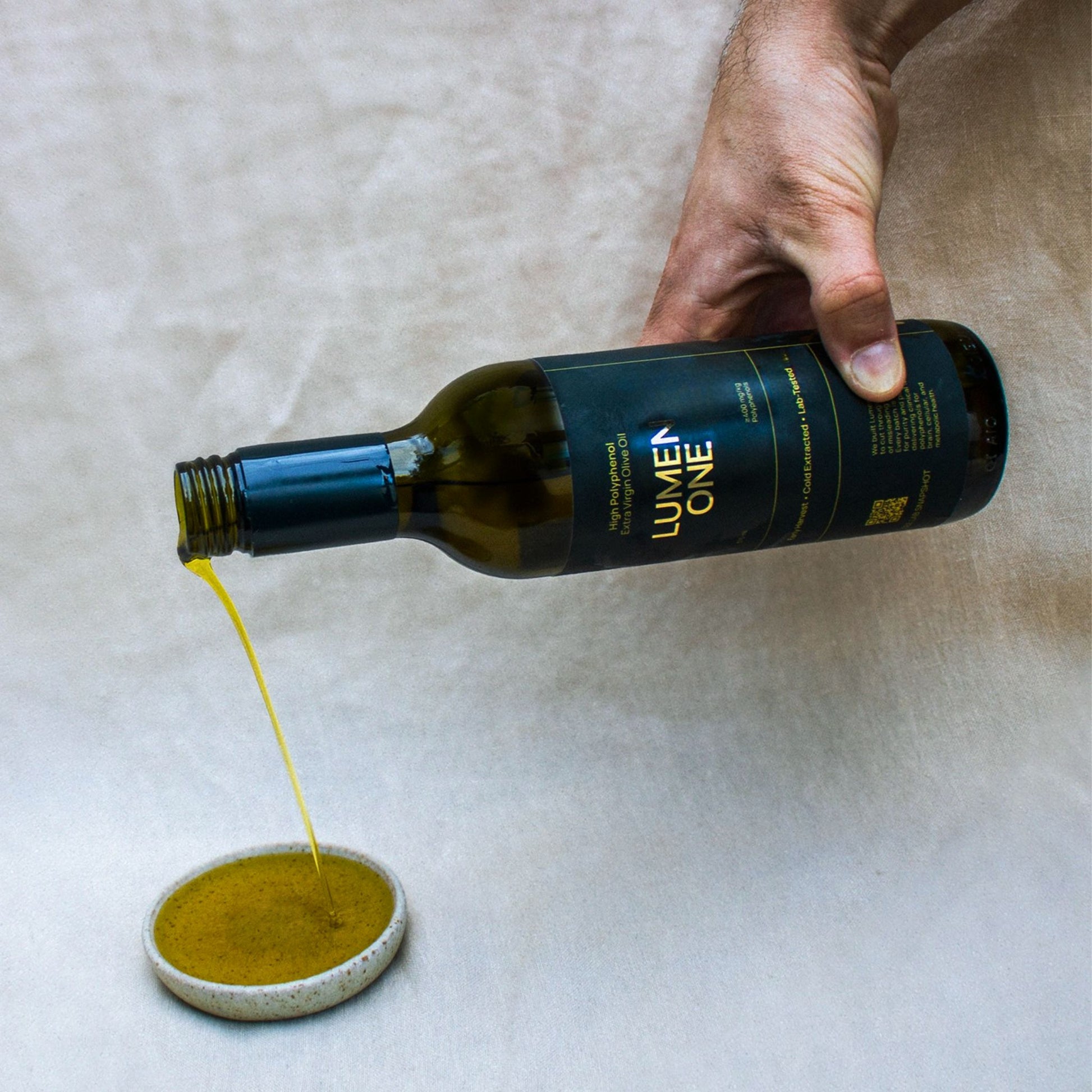 Hand pouring Lumen One high polyphenol olive oil onto a small dish on a textured surface.